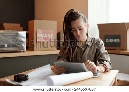 Young serious female solopreneur with tablet looking through online issues on screen while sitting by workplace in cafe under refit Royalty-Free Stock Photo #2445597087