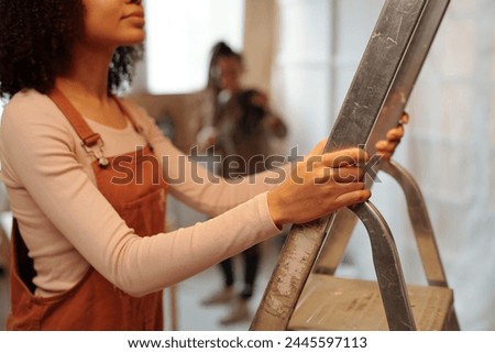 Cropped shot of young African American woman in long sleeve pullover and brown overalls holding by metallic stepladder during refit work Royalty-Free Stock Photo #2445597113