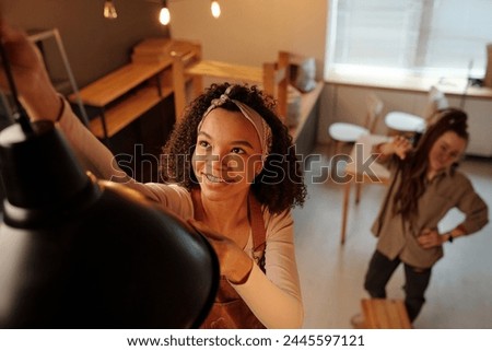 Young smiling African American female owner of new cafe hanging lamp over table during refit and renovation works while colleague helping her Royalty-Free Stock Photo #2445597121