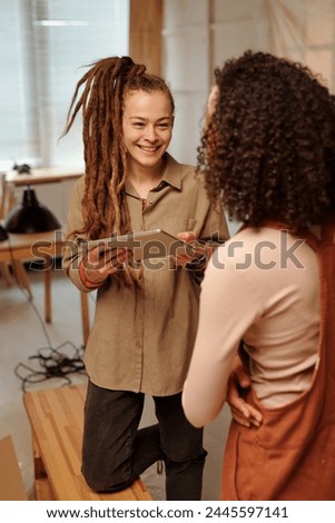 Young smiling businesswoman with tablet looking at her colleague during discussion of working points while standing in cafe under refit Royalty-Free Stock Photo #2445597141