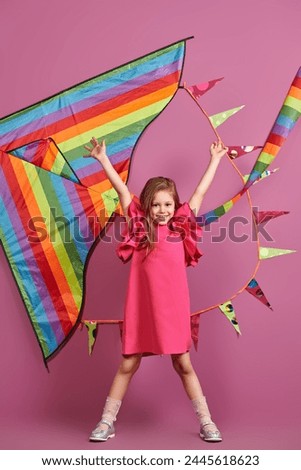 Dreams and imagination. Summer holidays. Happy cute girl in a bright fuchsia dress posing cheerfully against a rainbow kite and pink background. Children's fashion.