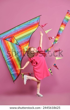 Summer holidays time. Happy cute girl in a bright fuchsia dress posing cheerfully against a rainbow kite and pink background. Children's fashion. Dreams and imagination.