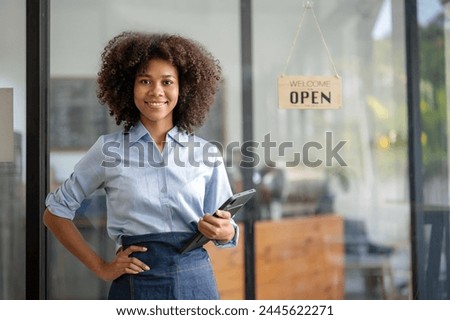 Female African coffee shop small business owner wearing apron standing  and in the background there is a welcome sign