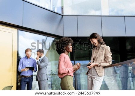 Two professional women chat animatedly, while male colleagues discuss in the background.