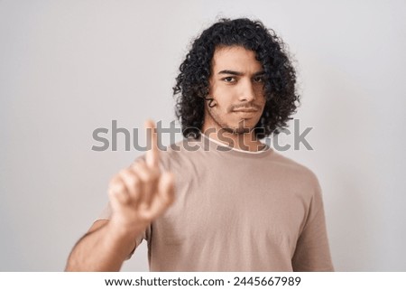 Hispanic man with curly hair standing over white background pointing with finger up and angry expression, showing no gesture 