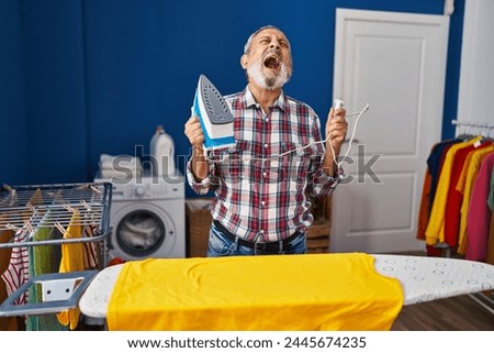Enraged senior man in laundry room, furious, screaming, iron machine in hand. frantic, frustrated and serious expression of anger, shouting at the housework. Royalty-Free Stock Photo #2445674235