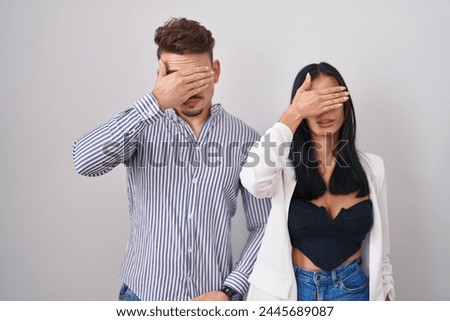 Young hispanic couple standing over white background covering eyes with hand, looking serious and sad. sightless, hiding and rejection concept  Royalty-Free Stock Photo #2445689087
