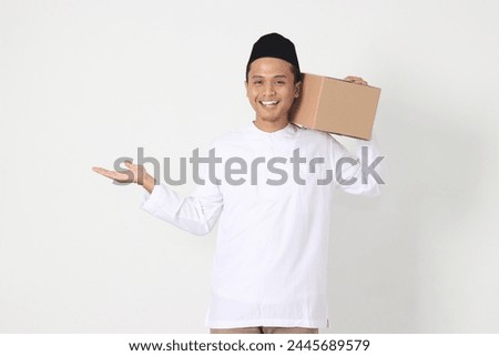 Portrait of attractive Asian muslim man in koko shirt with peci pointing to the side while carrying cardboard box. Going home for Eid Mubarak. Isolated image on white background