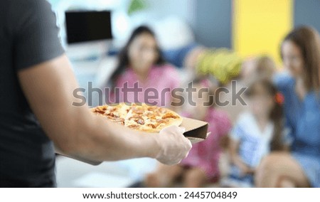 Male hands carry pizza on cardboard stand. In background, two women and two children sit on sofa.