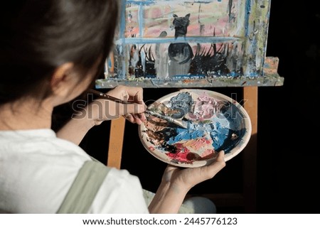 Young Asian artist woman holding a palette of colorful paints is intently painting acrylic paints on canvas in a painting studio.