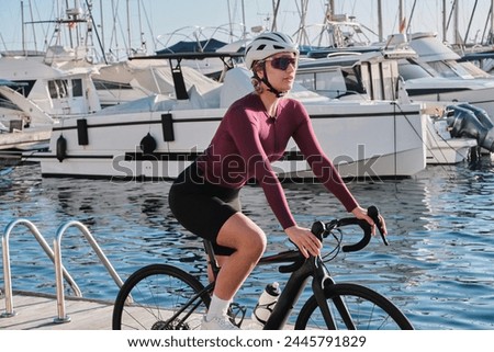 Woman cyclist wearing cycling kit and helmet riding a bicycle on the yacht club background. Road cycling concept. Female cyclist is training an E-bike. Woman cycling at sunset. Calp, Alicante, Spain.