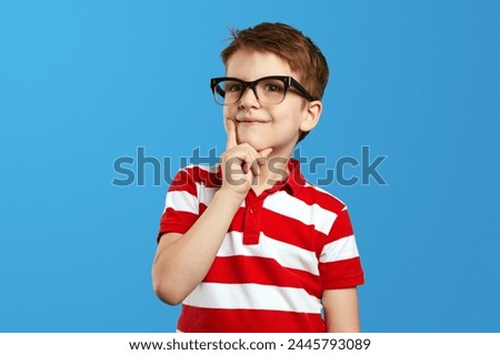 Smart thoughtful preschool boy in nerdy glasses and red striped polo shirt touching cheek and looking at camera while thinking against blue background.