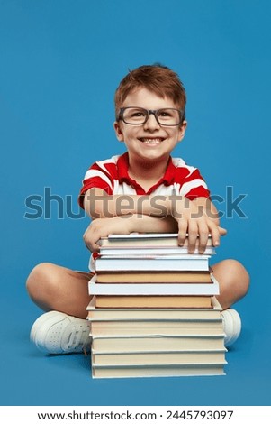Vertical photo of happy smart pupil boy with nerdy eyeglasses holding hands on bunch of books while looking at camera, wearing red striped shirt, isolated over blue background.