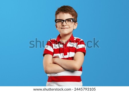 Smart little boy with wearing nerdy eyeglasses and striped red shirt smiling for camera while keeping hands crossed, standing isolated over blue background