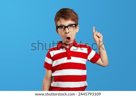 Adorable boy in nerdy eyeglasses and red striped shirt opening mouth and pointing up while having idea against blue background. Back to school concept