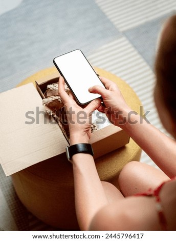 Female person customer of internet store unpacking online order and taking a photo of goods using smartphone with blank screen.