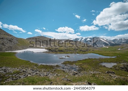 Beautiful small lake with snowfield among green grassy hills and rocks with view to snow-capped mountain range. Scenic landscape with alpine lake and snowy high mountains. Mountain lake in highlands. Royalty-Free Stock Photo #2445804379
