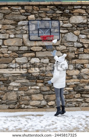 A woman in winter attire playing with a blue basketball against a stone wall with a hoop during a snowy day