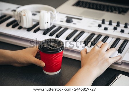 A woman musician plays the keyboard of a synthesizer and holds a red cup of coffee in her hand at her workplace. Royalty-Free Stock Photo #2445819729