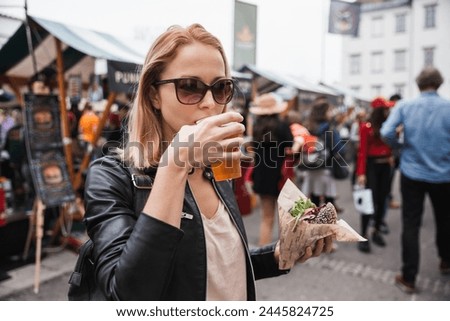 Beautiful young woman holding delicious organic salmon vegetarian burger and drinking homebrewed IPA beer on open air beer an burger urban street food festival in Ljubljana, Slovenia Royalty-Free Stock Photo #2445824725