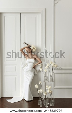 Chic and charming lady of European origin, featuring a polished makeup look, clad in a white luxurious dress, holding a bouquet of white roses, radiating elegance in a refined white studio.