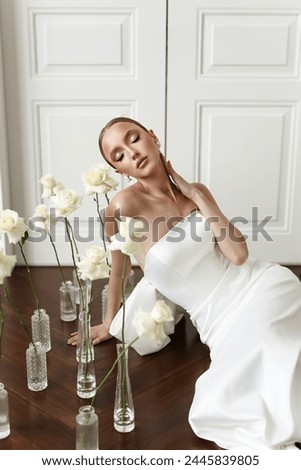 A vision of elegance, the young woman in her stunning white dress and immaculate makeup strikes a pose amidst the artful arrangement of white roses, creating a picture-perfect composition.