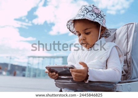 Cute baby child watching cartoons with smartphone, sitting at baby stroller outdoors.