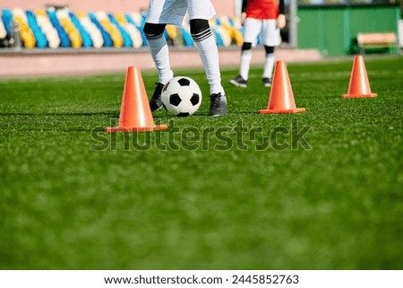 soccer player is deftly kicking a soccer ball through a series of orange cones set up in a training drill. The players focus, agility, and control are evident as they navigate the course with finesse. Royalty-Free Stock Photo #2445852763