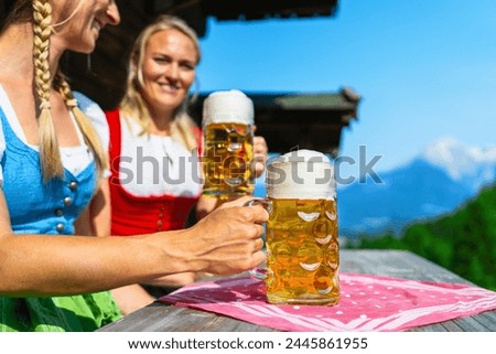 smiling german girls in traditional dirndls or tracht toasting beer mugs in front of the bavarian alpine mountains behind a alpine cabin. Oktoberfest concept image Royalty-Free Stock Photo #2445861955