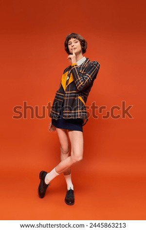 stylish student in checkered blazer and skirt showing hush on orange background, college uniform