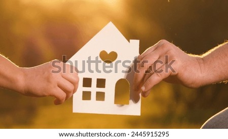 Woman and man hold cut-out cardboard house with hands on both sides. Parent with child grasp cardboard house each holding one side with hand. Father and mother clutch cardboard house dreaming of home Royalty-Free Stock Photo #2445915295