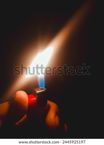 A close-up photo of a person's hand holding a lighter with a small flame burning in the dark. The flame illuminates the fingers and part of the lighter.