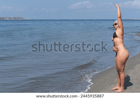 A woman in a swimsuit stretches on the shore before a swim, with the ocean behind her.