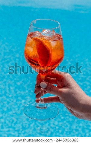 Aperol Spritz alcoholic cocktail in glass goblet in woman's hand with white nail polish on blue water pool background  Royalty-Free Stock Photo #2445936363