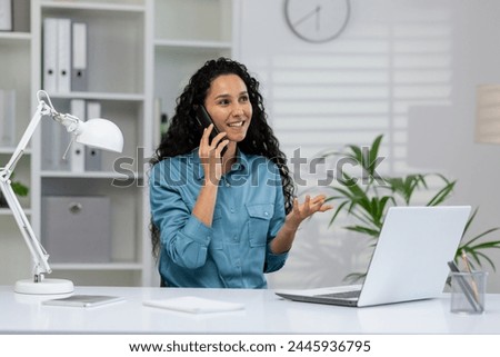 Cheerful businesswoman having a conversation on a smartphone with a laptop on her desk in a modern office setting.