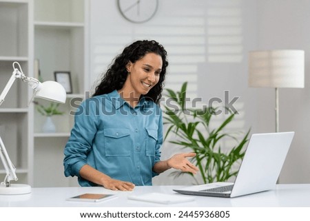 Friendly woman smiling and gesturing at the camera during a video call with a bright home office background.