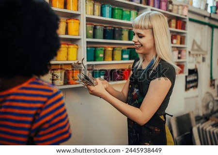 Smiling multicultural female graphic workers standing at printing shop with color swatch in hands and choosing paint buckets on shelves. Portrait of female technician with color swatch at facility.