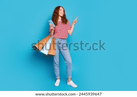 Full size photo of cute girl hold new clothes in bags smartphone look directing at discount empty space isolated on blue color background