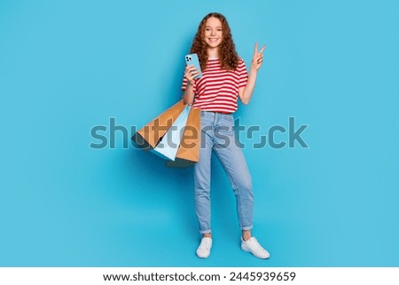 Full size photo of cheerful girl wear stylish t-shirt hold new clothes in bags smartphone show v-sign isolated on blue color background