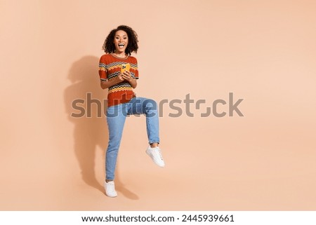 Full size photo of positive woman wear knit t-shirt denim pants hold smartphone stand on one leg isolated on beige color background