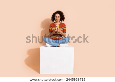 Full size photo of positive woman wear knit t-shirt sit on cube read notification on smartphone isolated on beige color background
