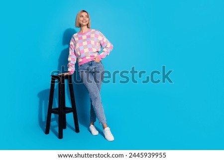 Full size photo of positive woman dressed colorful sweater lean on stool look at proposition empty space isolated on blue color background