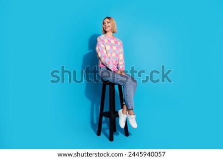 Full length photo of cheerful girl wear checkered pullover look at proposition empty space sit on chair isolated on blue color background