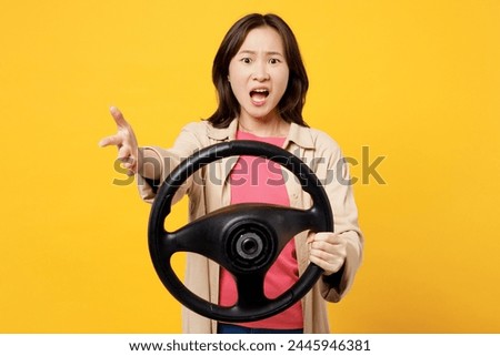 Young indignant woman of Asian ethnicity she wear pink t-shirt beige shirt pastel casual clothes hold steering wheel driving car isolated on plain yellow background studio portrait. Lifestyle portrait