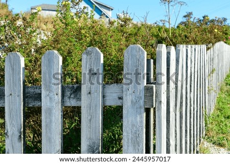 Corner of a wood picket fence with grey faded boards. The structure has been exposed to the weather elements with white paint peeling. Behind the barrier is a lush green shrub or hedge and a blue sky. Royalty-Free Stock Photo #2445957017