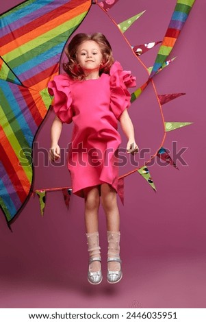 Happy cute girl in a bright fuchsia dress posing cheerfully against a rainbow kite and pink background. Children's fashion. Summer holidays. Dreams and imagination. Copy space.