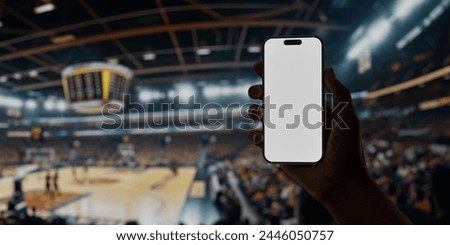 Black African-American man using smartphone with a blank screen during a game on basketball arena, perfect for showcasing apps or designs with a backdrop of live sports and enthusiastic fans