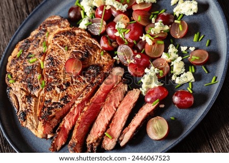 fried beef steak entrecote served with salad of red grape, crumbled blue mold cheese and chives on plate on dark wooden table, dutch angle view, close-up, Royalty-Free Stock Photo #2446067523