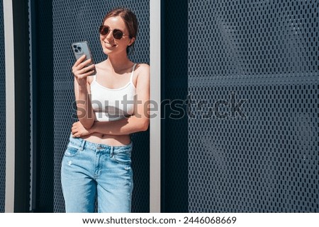 Young beautiful smiling hipster woman in trendy summer casual clothes. Carefree woman posing in the street. Positive model holds mobile phone, looks at cellphone screen, uses smartphone apps