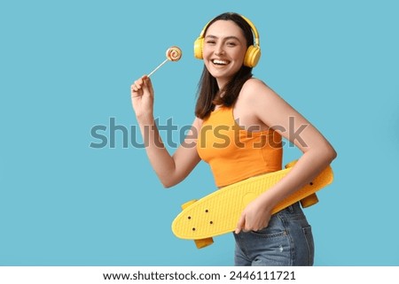 Beautiful young woman in headphones with sweet lollipop and skateboard on blue background